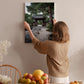 A woman hangs a framed picture of a Japanese garden on a wall in a room with a wooden chair, a fruit bowl, and a vase of flowers.