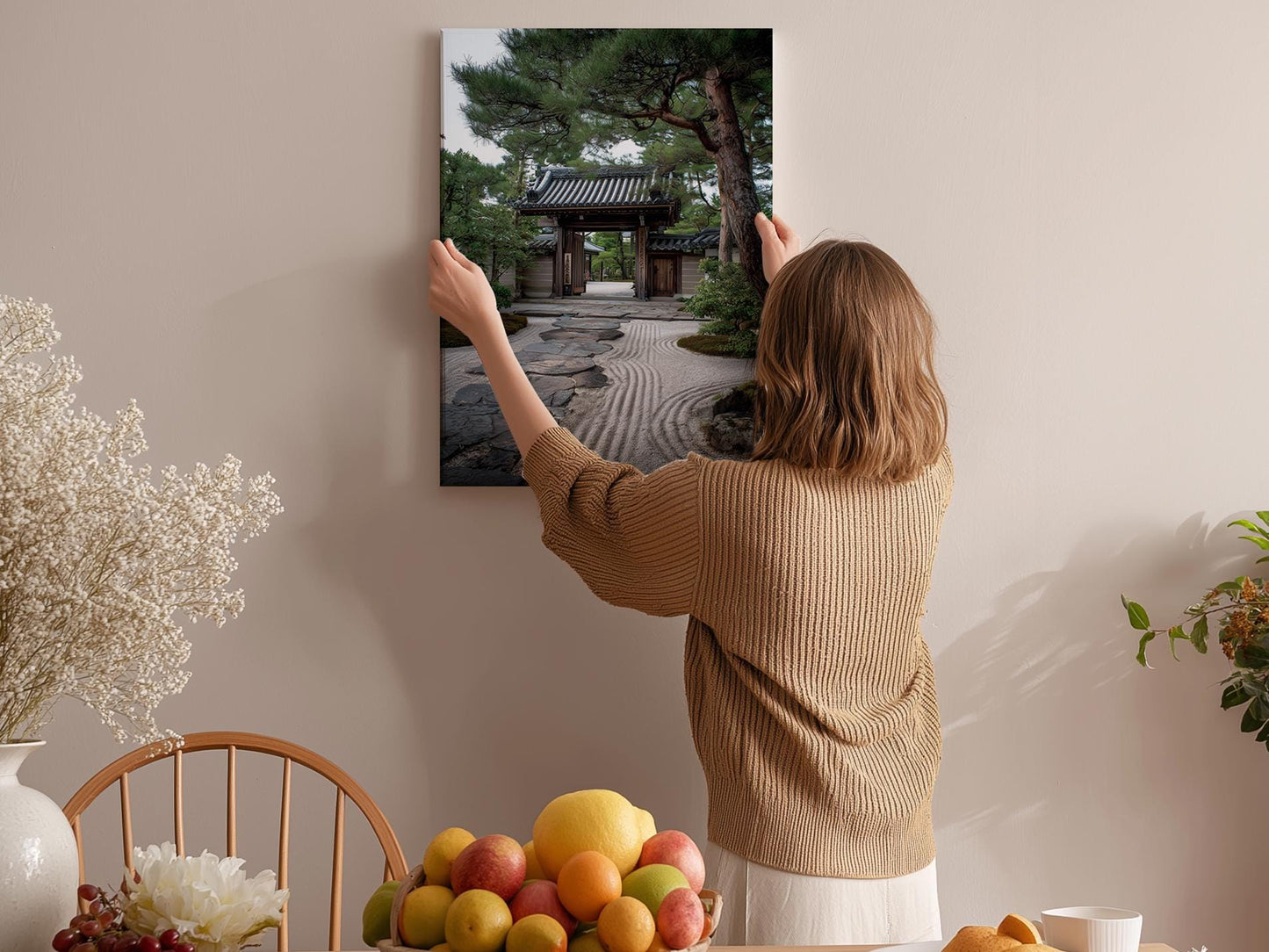 A woman hangs a framed picture of a Japanese garden on a wall in a room with a wooden chair, a fruit bowl, and a vase of flowers.