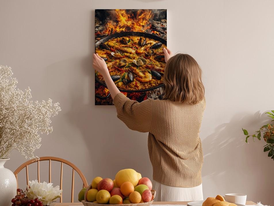 A woman hangs a framed picture of a seafood paella on a wall in a cozy kitchen setting.