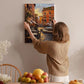A woman hangs a painting of a cityscape on a wall in a cozy room with a dining table, chairs, and various decorative items.
