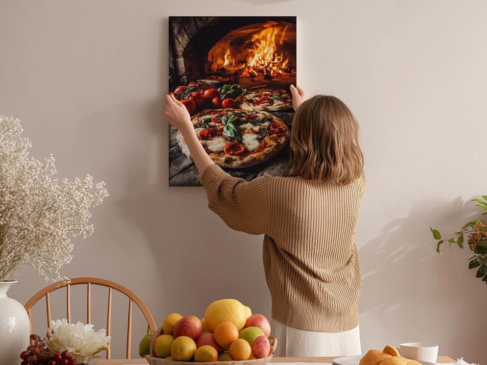 A woman hangs a framed picture of a pizza oven on a wall in a cozy kitchen setting.