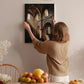 A woman hangs a framed picture on a wall in a room with a dining table, chairs, and various fruits and flowers.