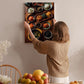 A woman hangs a framed picture of various bowls of food on a wall in a cozy kitchen setting.