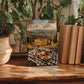 a wooden table with a framed photograph of a picnic scene, surrounded by potted plants and books.