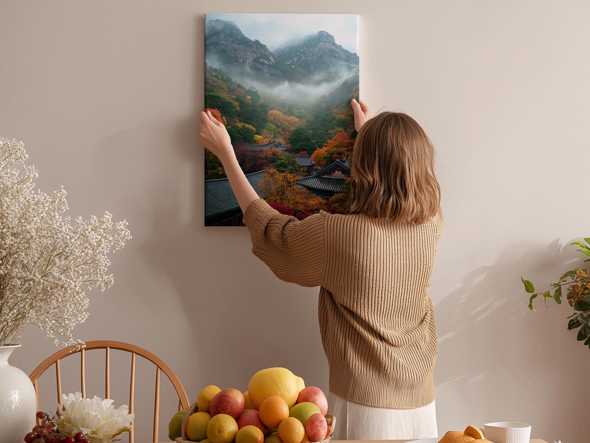A woman hangs a framed picture of a mountainous landscape on a wall in a cozy room with a dining table, chairs, and various fruits.
