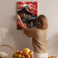 A woman hangs a framed picture of a traditional Japanese temple with red leaves on a wall in a room with a dining table, chairs, and various fruits.