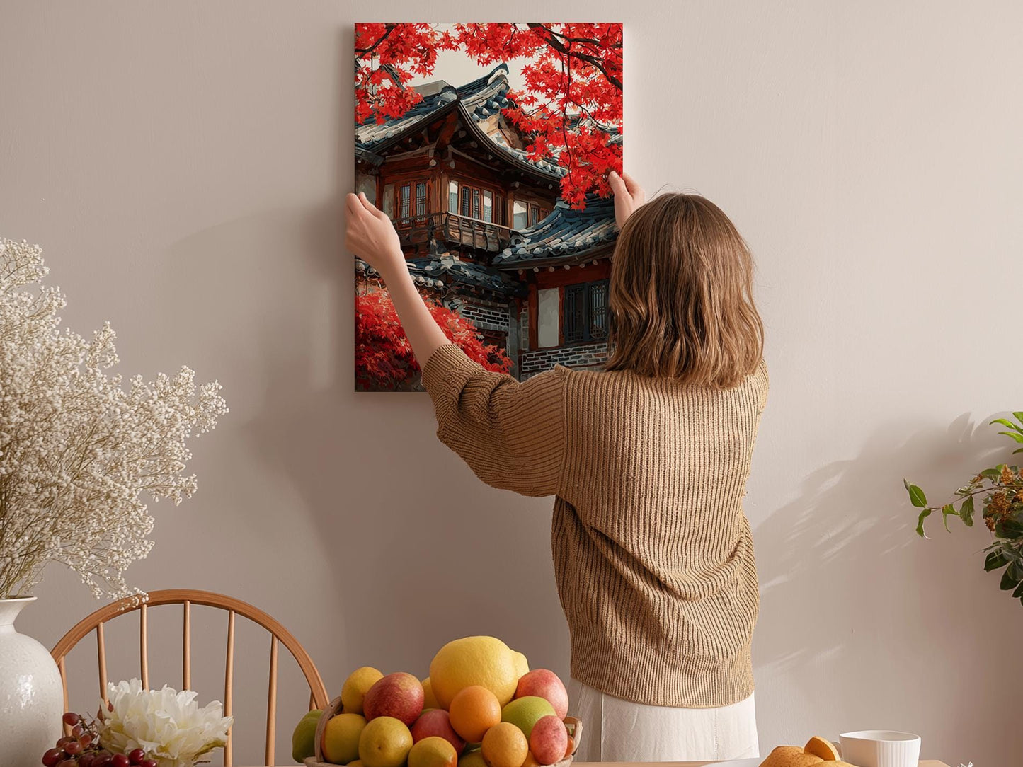 A woman hangs a framed picture of a traditional Japanese temple with red leaves on a wall in a room with a dining table, chairs, and various fruits.