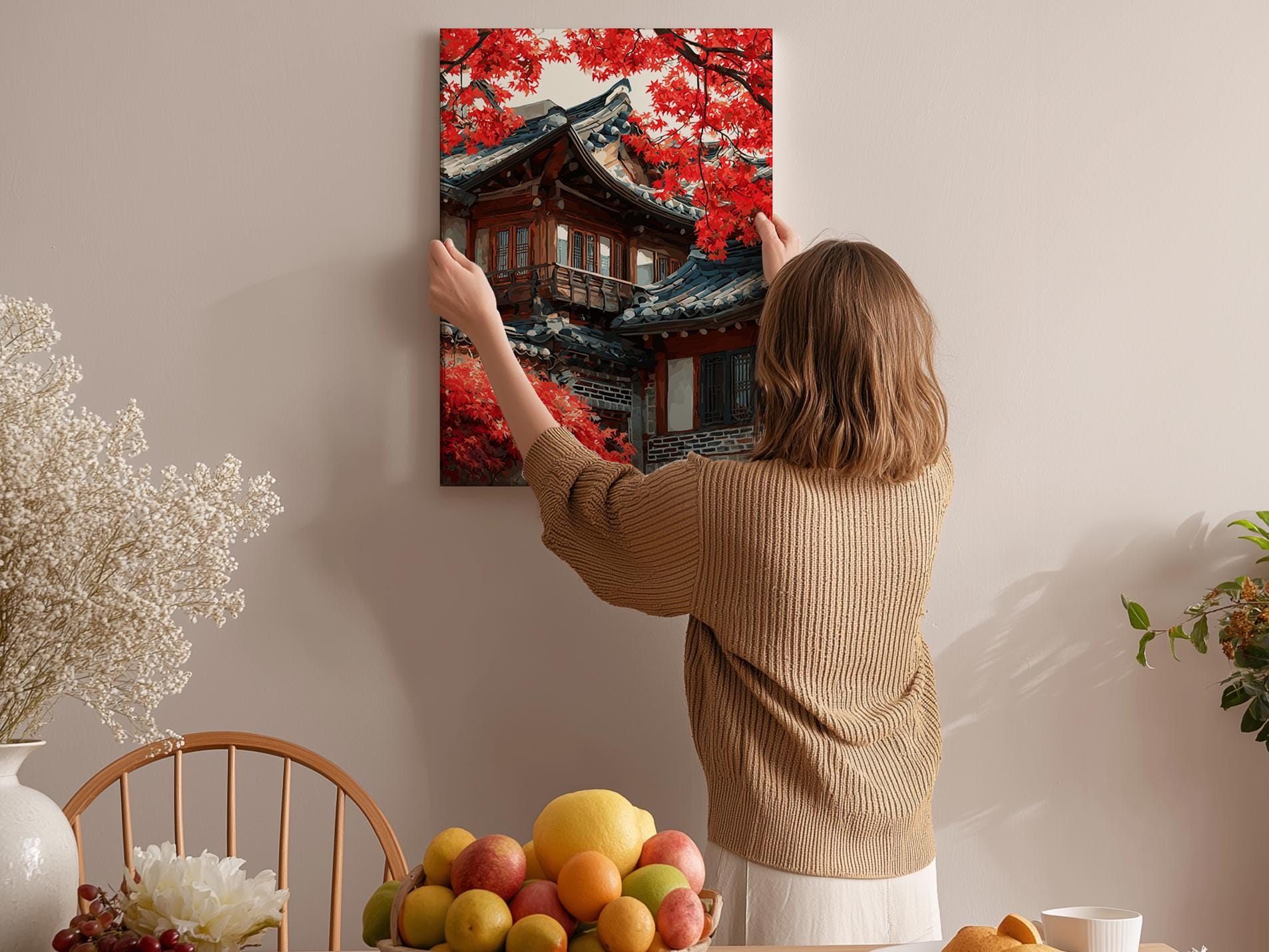 A woman hangs a framed picture of a traditional Japanese temple with red leaves on a wall in a room with a dining table, chairs, and various fruits.