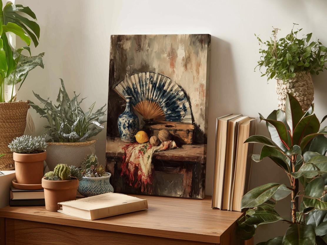 a wooden table with a painting of a still life scene, including a fan, fruit, and other decorative items. The table also has several books and potted plants, creating a cozy and artistic atmosphere.