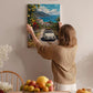 A woman hangs a framed painting of a car on a wall, surrounded by a cozy indoor setting with a dining table, chairs, and various fruits.