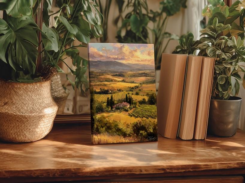 a wooden table with a framed landscape painting depicting a countryside scene, surrounded by potted plants and books.