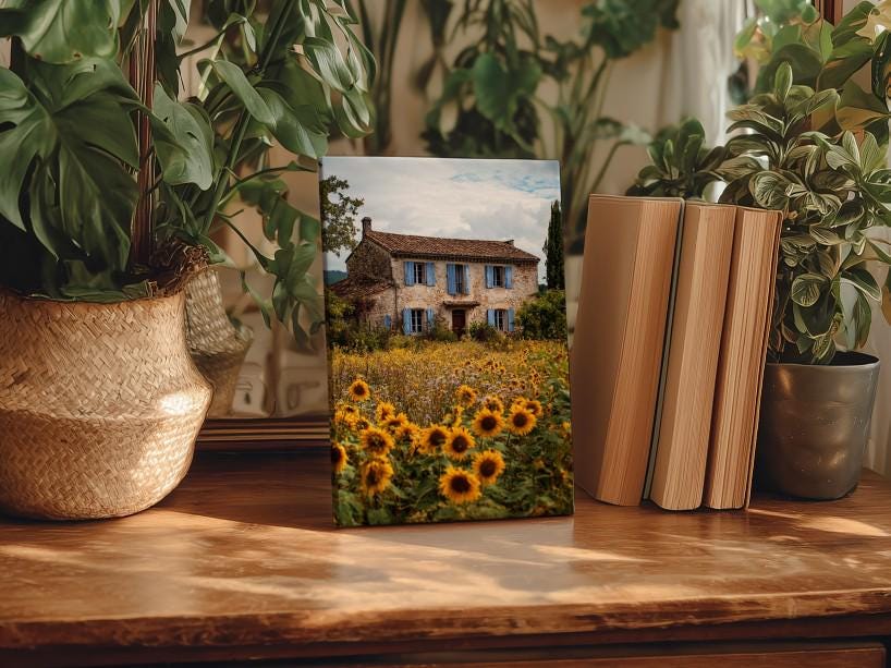 a wooden table with a framed photograph of a house surrounded by a field of sunflowers, placed in the center of the table. The table is also adorned with several potted plants, including a large potted plant on the left side and a smaller potted plant on the right side. There are also two books placed on the table, one on the right side and one on the left side.