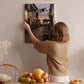 A woman hangs a framed picture of a cobblestone street with a sunset in the background on a wall in a cozy, homely setting.
