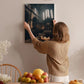 A woman hangs a framed photograph of a church interior on a wall in a room with a dining table, chairs, and various decorative items.