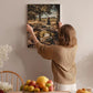 A woman hangs a framed picture of a picnic scene on a wall, surrounded by a table with fruit and flowers.