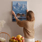 A woman hangs a painting of a mountain landscape on a wall in a cozy room with a dining table, chairs, and various decorative items.