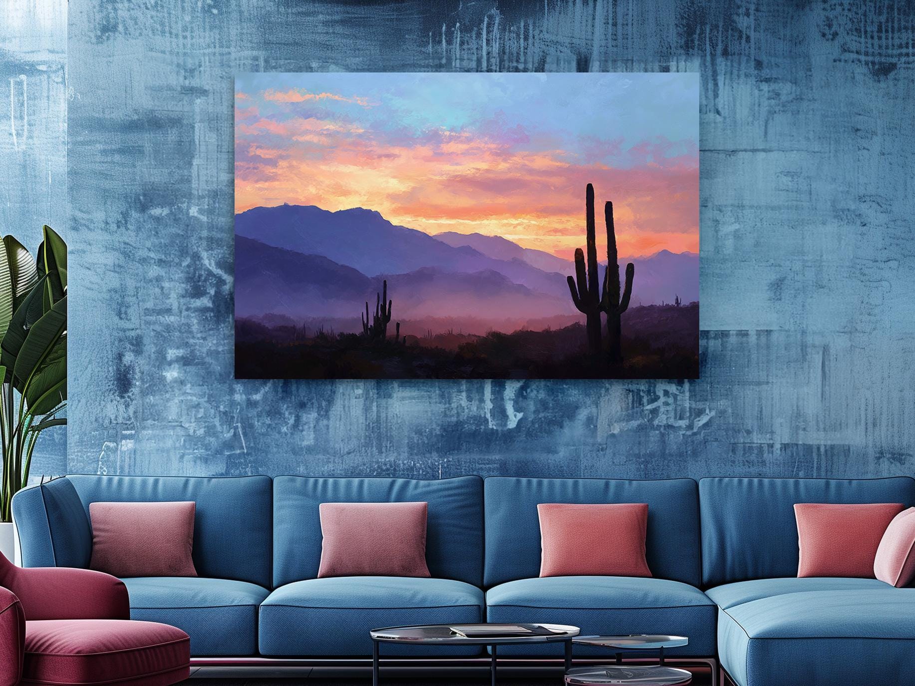 a large blue sofa with red throw pillows, positioned in front of a wall with a framed painting depicting a desert landscape with mountains and a cactus in the foreground.