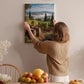 A woman hangs a framed picture of a vineyard on a wall in a cozy room with a dining table, chairs, and various fruits and flowers.