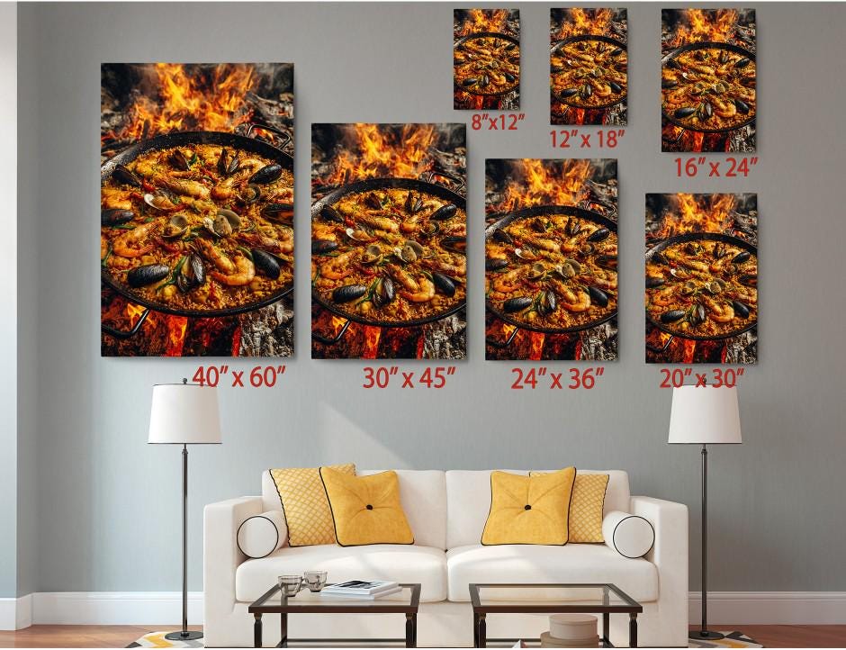 a living room with a white sofa and a coffee table. Above the sofa, there are several framed prints of the same image, depicting a large pan of seafood on a fire. The prints vary in size, ranging from 6 x 12&quot; to 24 x 36&quot;. The prints are arranged in a grid-like pattern on the wall, creating a visually appealing display.
