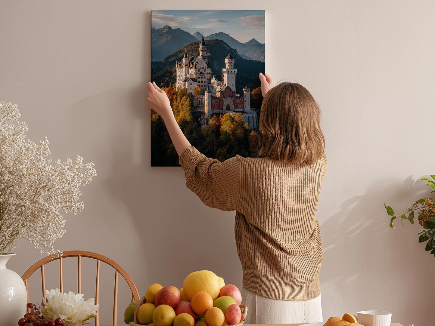 A woman hangs a framed picture of a castle on a wall in a cozy room with a dining table, chairs, and various decorative items.