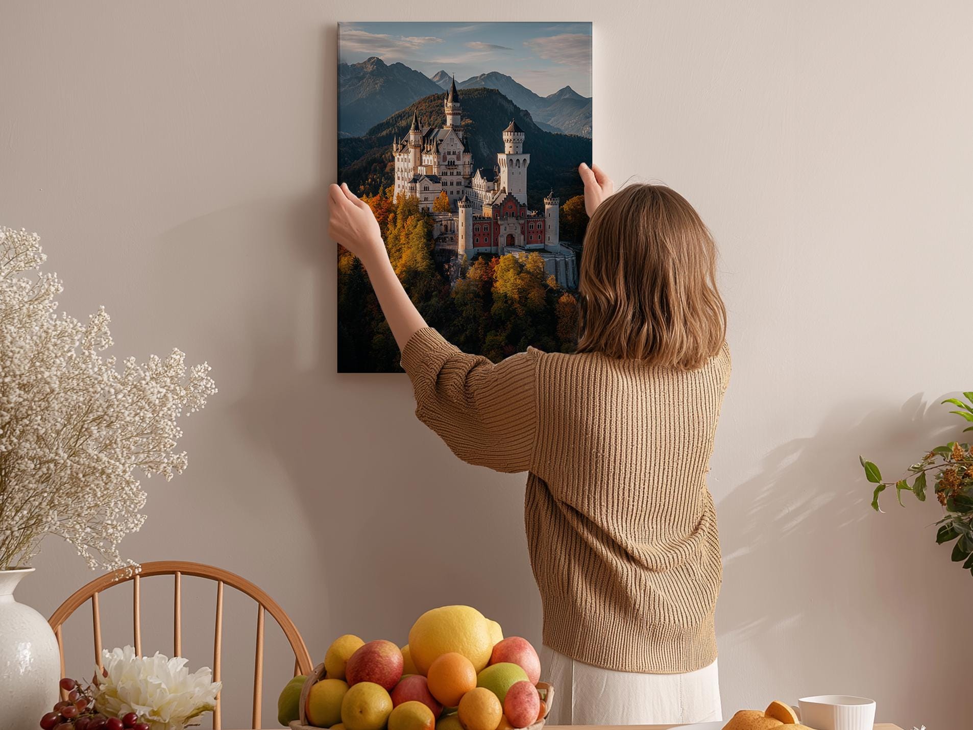 A woman hangs a framed picture of a castle on a wall in a cozy room with a dining table, chairs, and various decorative items.
