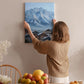 A woman hangs a painting of a mountain village on a wall in a cozy room with a dining table, chairs, and various fruits.