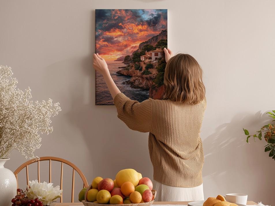A woman hangs a framed picture of a coastal landscape on a wall, surrounded by a dining table with fruit and flowers.