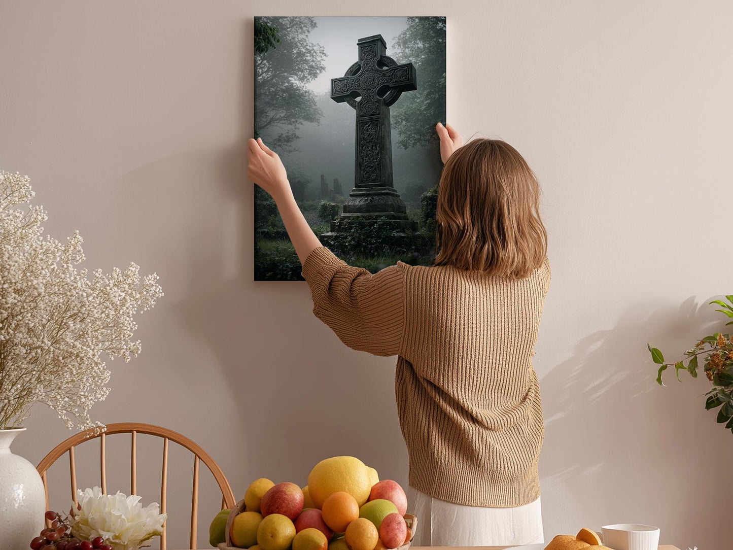 A woman hangs a framed image of a cross on a wall, with a table of fruit and flowers in the foreground.