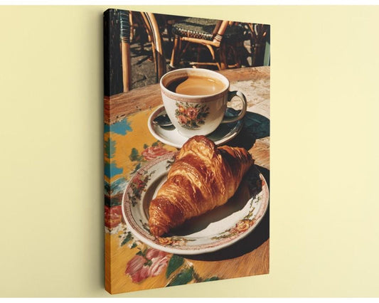 A croissant on a floral plate and a cup of coffee on a wooden table, with a wicker chair visible in the background.