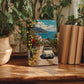 a wooden table with a framed painting of a car driving down a coastal road, surrounded by lush greenery and a scenic view of the sea.