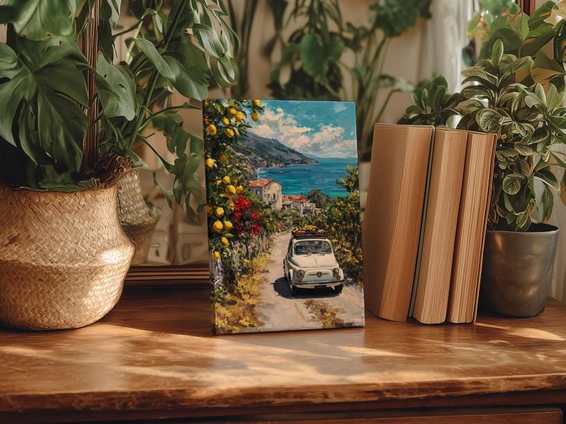 a wooden table with a framed painting of a car driving down a coastal road, surrounded by lush greenery and a scenic view of the sea.