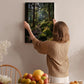 A woman hangs a framed picture of a forest on a wall in a cozy room with a dining table, chairs, and various fruits.