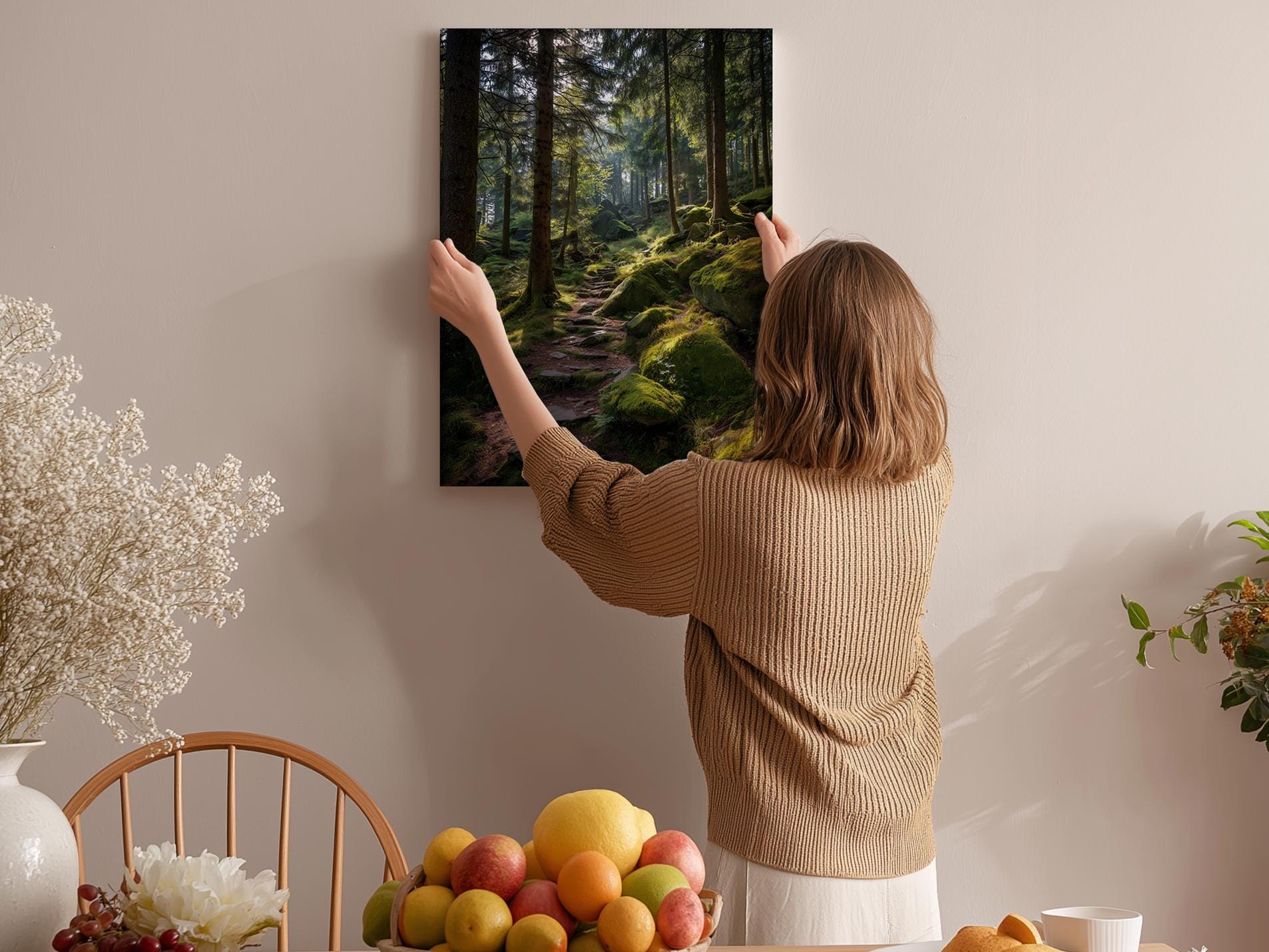 A woman hangs a framed picture of a forest on a wall in a cozy room with a dining table, chairs, and various fruits.
