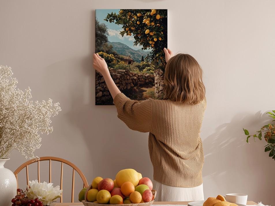 A woman hangs a framed picture of an orange grove on a wall in a cozy room with a dining table, chairs, and various fruits.