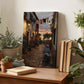 a cozy indoor setting with a wooden table displaying various potted plants, a book, and a framed picture of a cobblestone street at sunset.