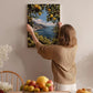 A woman hangs a framed picture of a scenic landscape on a wall, surrounded by a dining table with fruit and flowers.