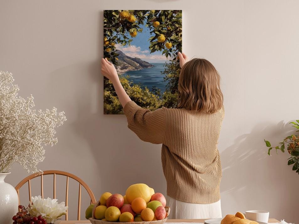 A woman hangs a framed picture of a scenic landscape on a wall, surrounded by a dining table with fruit and flowers.