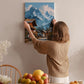 A woman hangs a painting of a mountain village on a wall in a cozy room with a dining table, chairs, and various fruits.