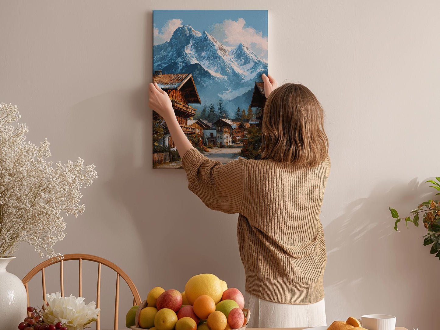 A woman hangs a painting of a mountain village on a wall in a cozy room with a dining table, chairs, and various fruits.