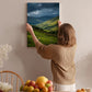 A woman hangs a landscape painting on a wall, which depicts a countryside scene with rolling hills and a valley.