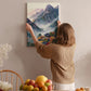 A woman hangs a painting of a mountain landscape on a wall in a cozy room with a dining table, chairs, and various fruits.