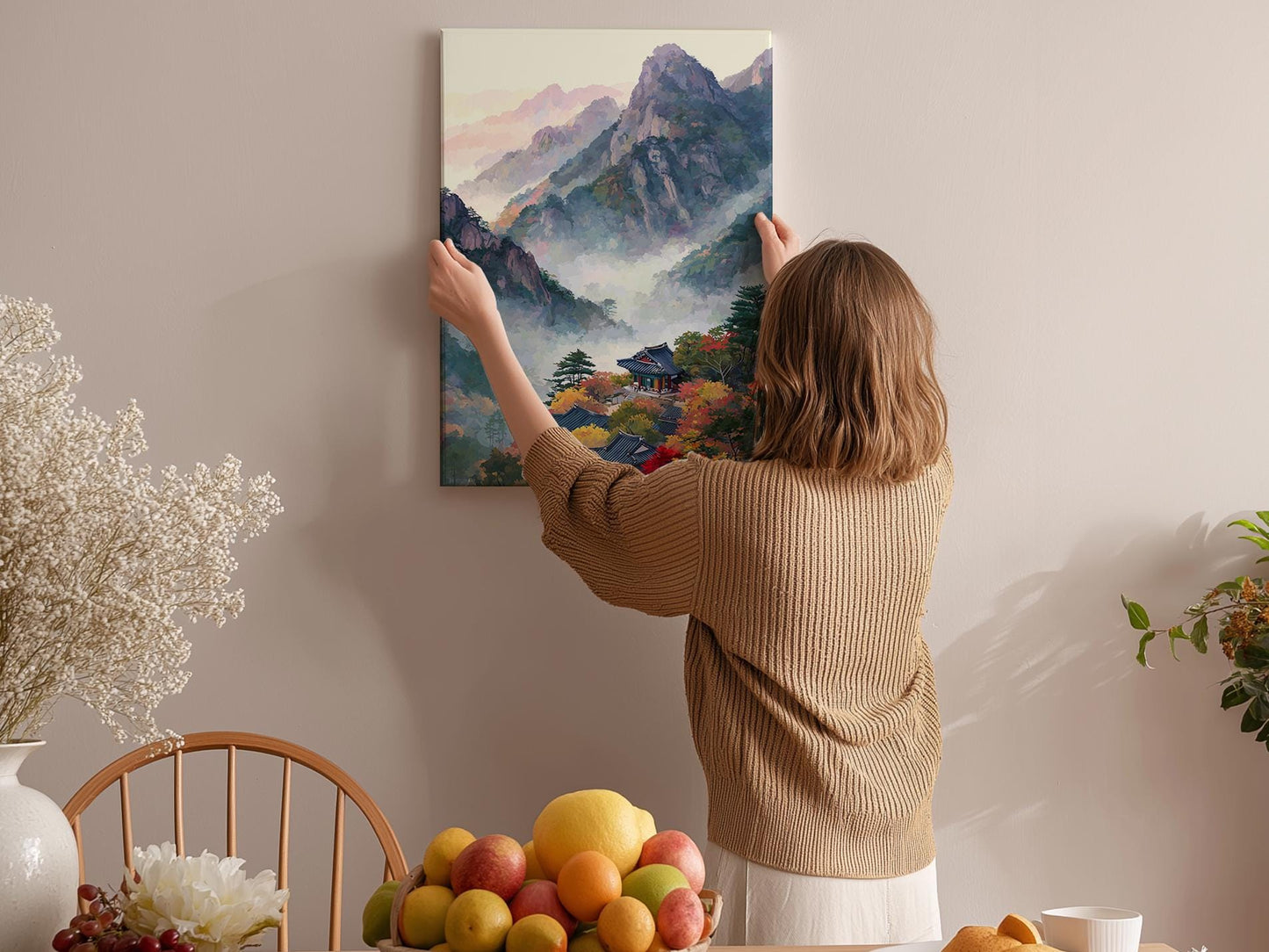 A woman hangs a painting of a mountain landscape on a wall in a cozy room with a dining table, chairs, and various fruits.