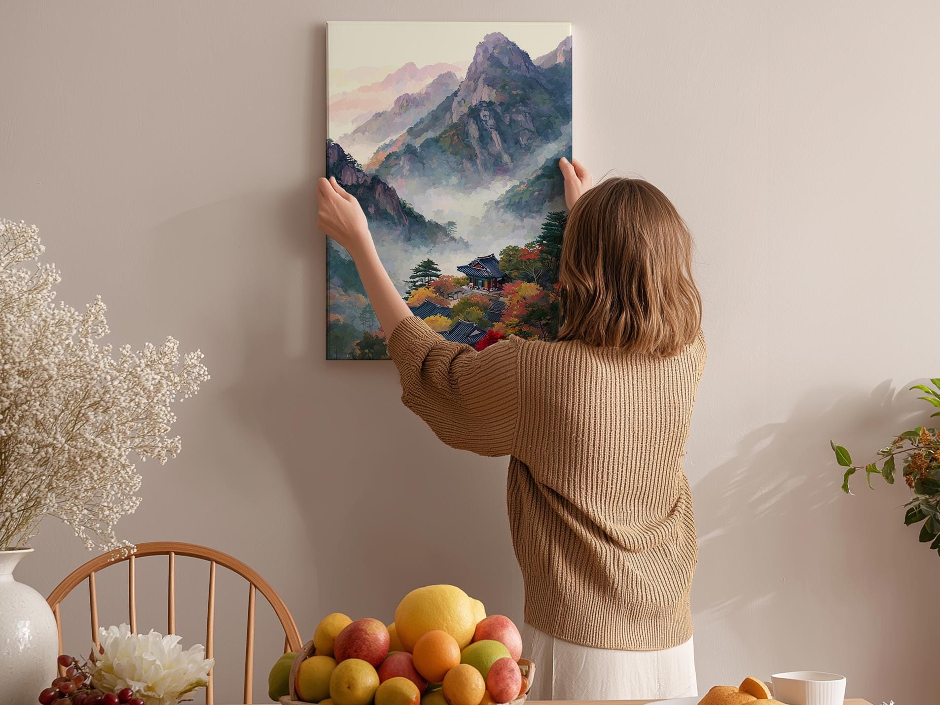 A woman hangs a painting of a mountain landscape on a wall in a cozy room with a dining table, chairs, and various fruits.
