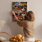 A woman hangs a framed photograph of a fruit stand on a wall in a room with a dining table, chairs, and various decorative items.
