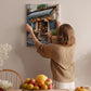 A woman hangs a framed picture of a fruit stand on a wall in a cozy kitchen setting.
