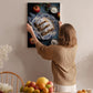 A woman hangs a framed photograph of a dessert on a wall, surrounded by various fruits and flowers in a cozy, homely setting.