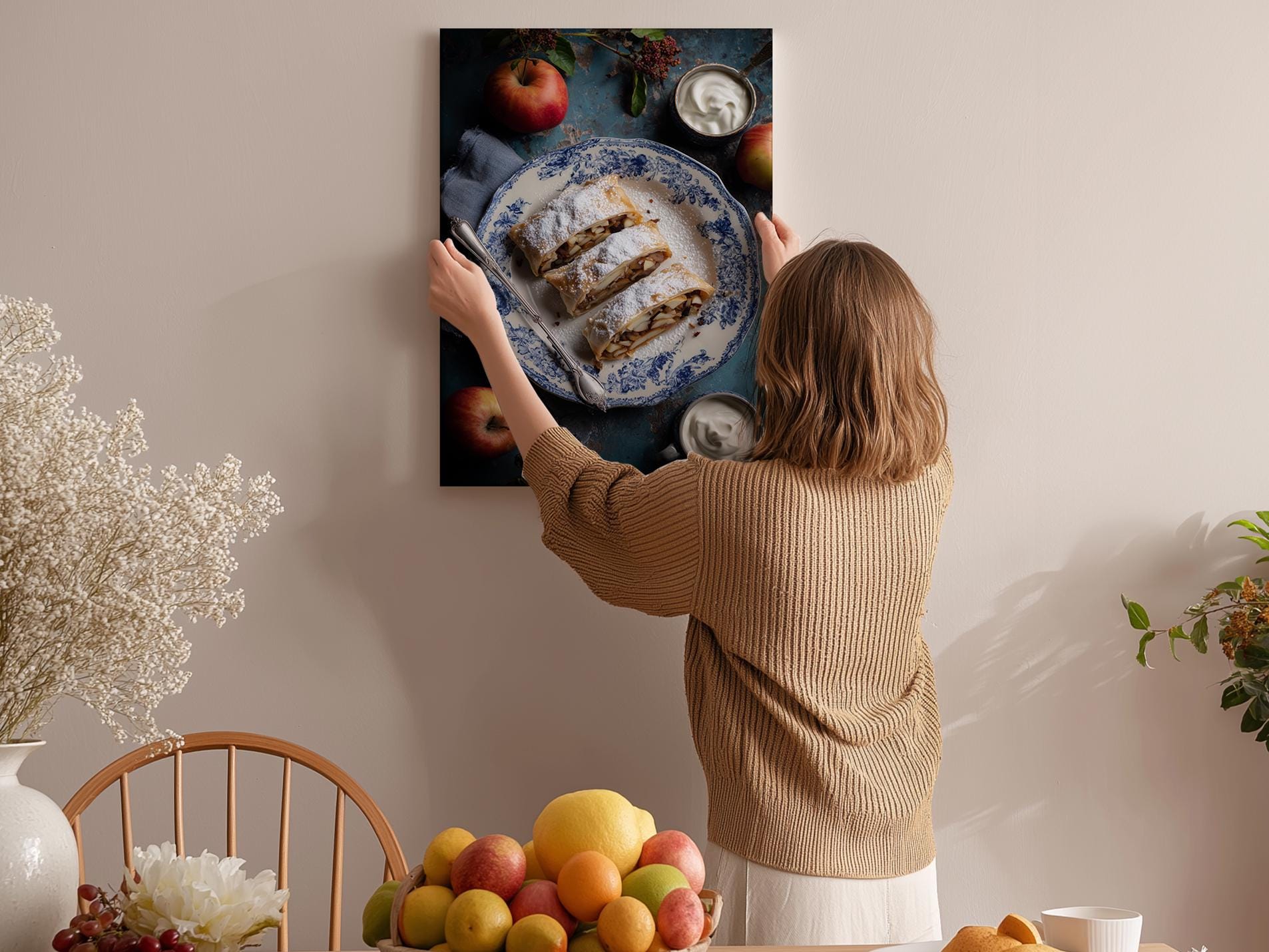 A woman hangs a framed photograph of a dessert on a wall, surrounded by various fruits and flowers in a cozy, homely setting.