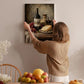A woman hangs a framed painting of a still life of wine, cheese, and fruit on a wall in a cozy, homely setting.