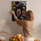 A woman hangs a framed picture of a fruit platter on a wall in a cozy kitchen setting.