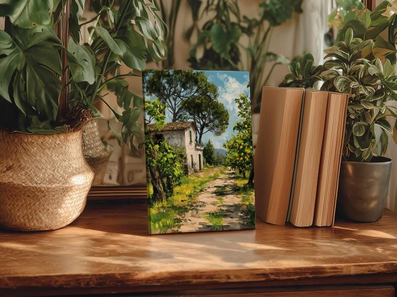 a painting of a house on a path surrounded by trees, displayed on a wooden surface. The painting is accompanied by a stack of books and several potted plants, creating a cozy and artistic atmosphere.